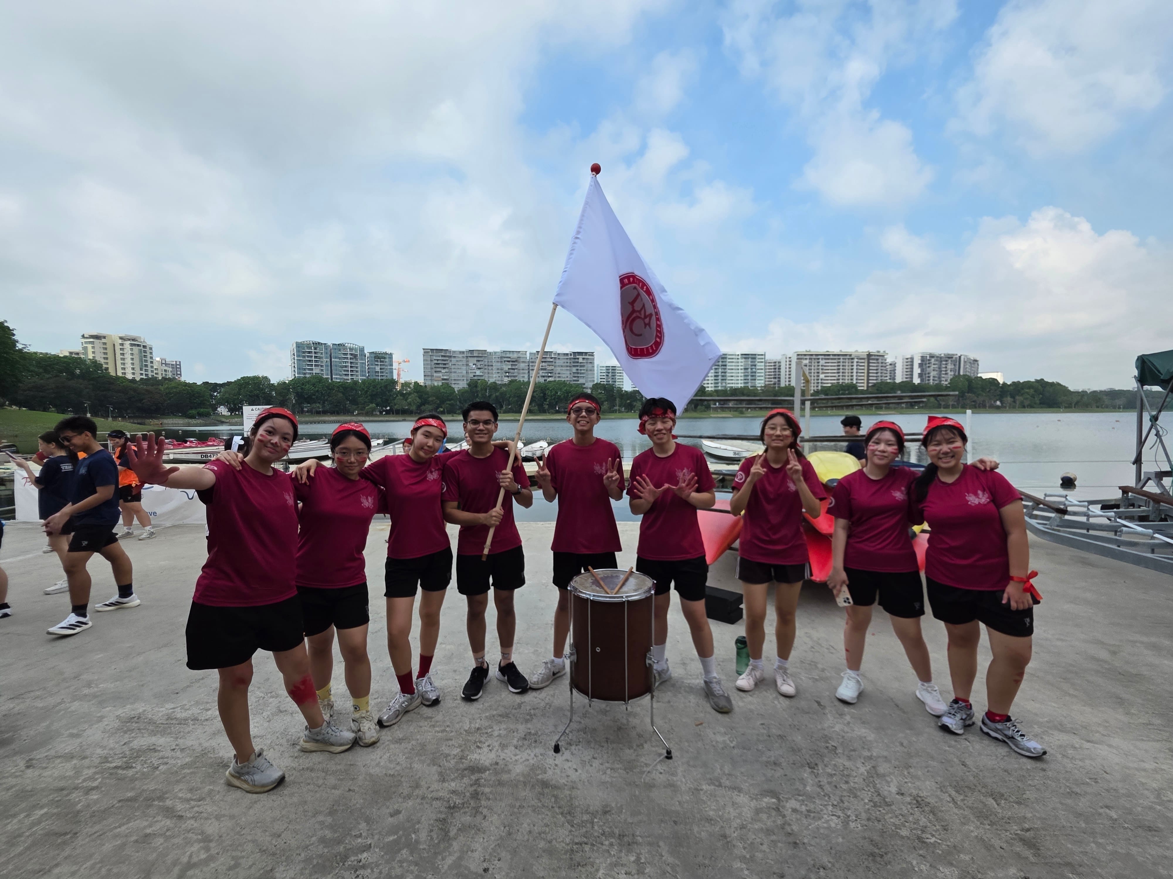 Nine students in maroon Aquila house T-shirts and red headbands pose by a waterfront, holding their house flag and a drum, smiling energetically.